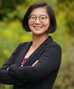 Headshot of Greta Cheng. Standing in front of green foliage, arms crossed.